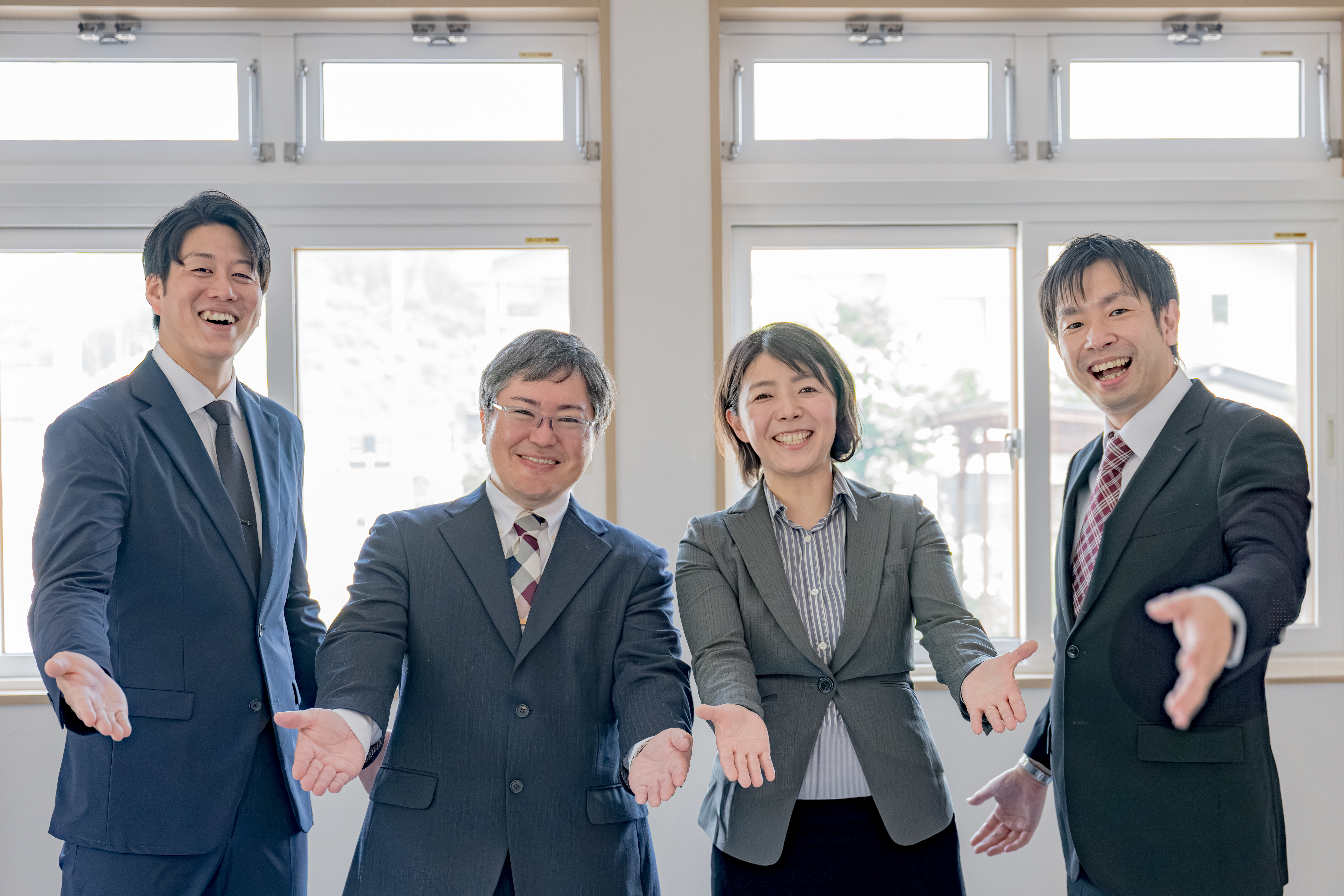 明るい室内で、4人のスーツ姿のビジネスパーソンが並んで立ち、笑顔で両手を広げ、歓迎や招き入れるジェスチャーをしている。左から背の高い男性、メガネをかけた男性、女性、そして右端の男性。背景には大きな窓がある。
