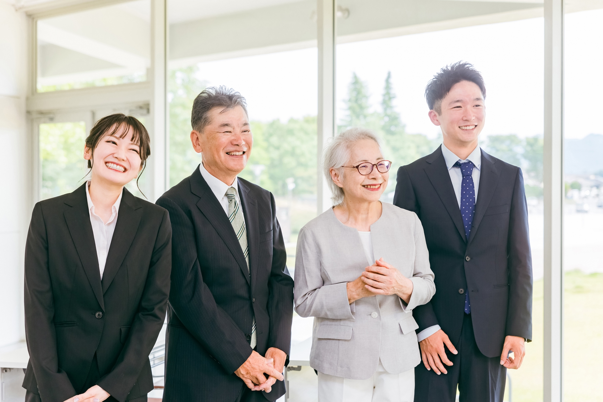 明るい室内の大きな窓際で、横一列に並んで立つ4人の日本人ビジネスパーソンの上半身写真。左から順に、黒いリクルートスーツと白のシャツを着用し頭を左に傾けて笑顔を見せる若い女性。黒のストライプ柄スーツと白のワイシャツ、ストライプ柄のネクタイを着用し正面右側を向いて笑うシニア男性。グレーのジャケットと白のインナーを着用し眼鏡をかけて胸の前で両手を組むシニア女性。ネイビーのスーツと薄いブルーのワイシャツ、ドット柄のネクタイを着用して正面を向いて笑う若い男性。背景は全面ガラス張りの大きな窓で、屋外の緑の木々や白い建物、青空がぼかして写り込んでいる。