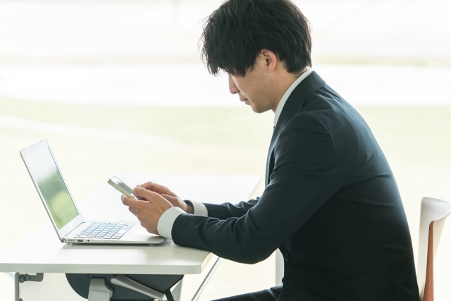 窓際のデスクでノートパソコンを開き、スマートフォンを手に画面を確認するスーツ姿の若い男性ビジネスパーソンの横顔。