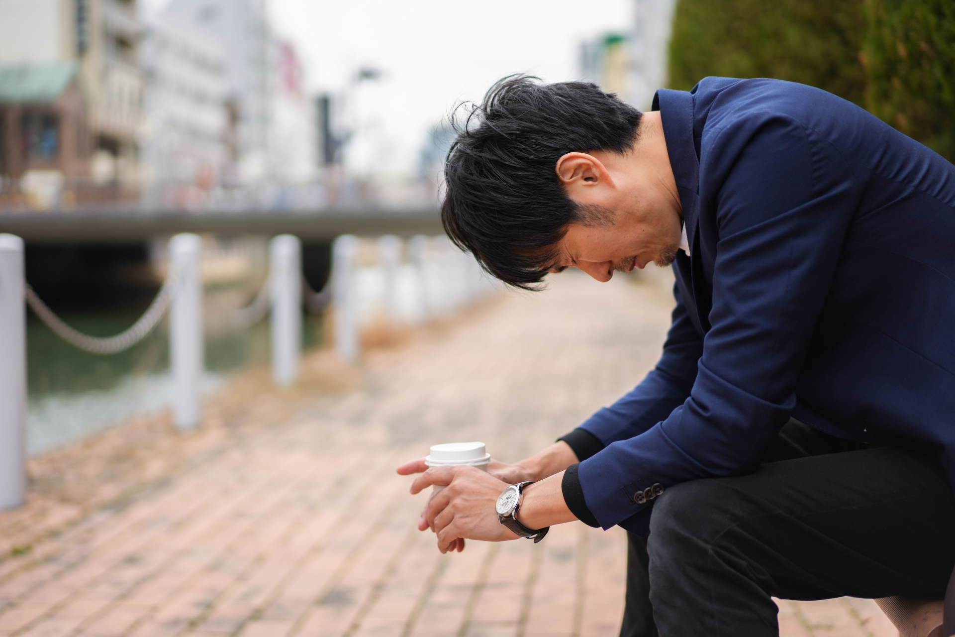 川沿いの歩道に設置されたベンチに座る、スーツを着用した男性の画像。男性は両手でカップを包み込むように持ち、頭を下げて下を向いている。背景には川と橋、市街地の建物が写り込んでいる。全体として、思索にふけっているような、あるいは疲労や悩みを抱えているかのような、静かで落ち着いた雰囲気の光景である。