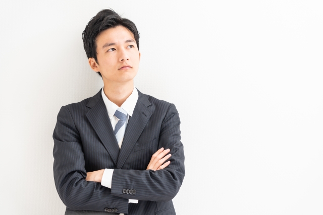 白い壁面を背景に、スーツを着用して腕組みをし、斜め右上を見上げて考え込む若い男性ビジネスパーソンの上半身写真。男性は黒のストライプ柄スーツジャケット、白いワイシャツ、そしてライトブルーと白のストライプ柄ネクタイを着用している。胸の前で両腕を深く組み、口元を引き締めて真剣な表情を浮かべながら、視線を右斜め上方に向けている。