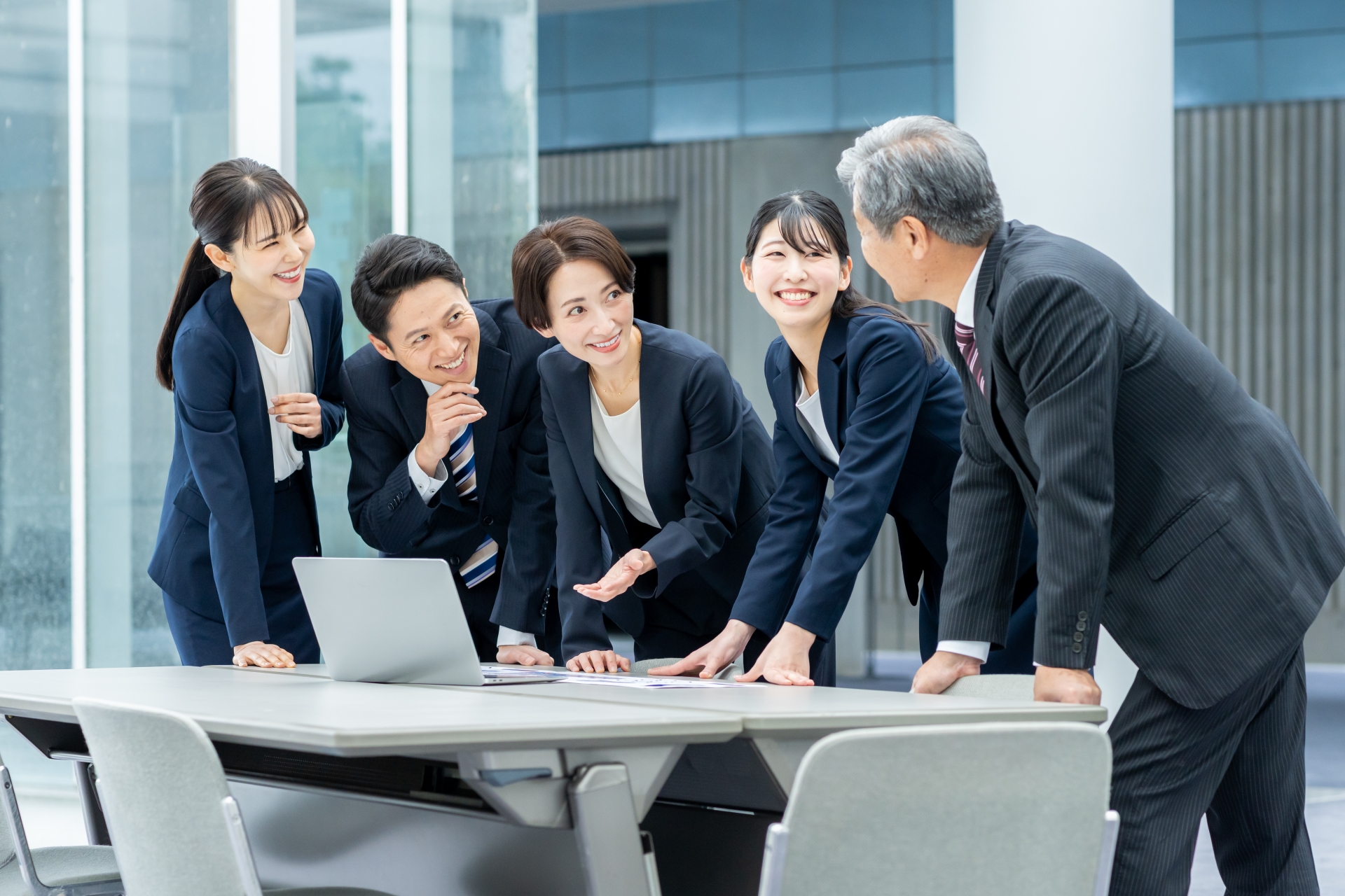 明るいオフィス（または近代的なビル内の会議スペース）で、1台の長机に置かれたノートパソコンと書類を囲み、立ってディスカッションを行う5人の日本人ビジネスパーソン。机の左側にはオフィスカジュアルやネイビーのスーツを着用した女性2人と男性1人が並び、中央の女性が左手をノートパソコンに向けて差し出している。机の右側にはネイビーのスーツを着用した若い女性、そして手前に背を向ける形で立つストライプ柄スーツのシニア男性が向かい合っており、全員が笑顔で中央のノートパソコンや書類を見つめている。背景には、大きなガラス窓から差し込む外光と、薄くぼかされた近代的な建物の一部が写り込んでいる。