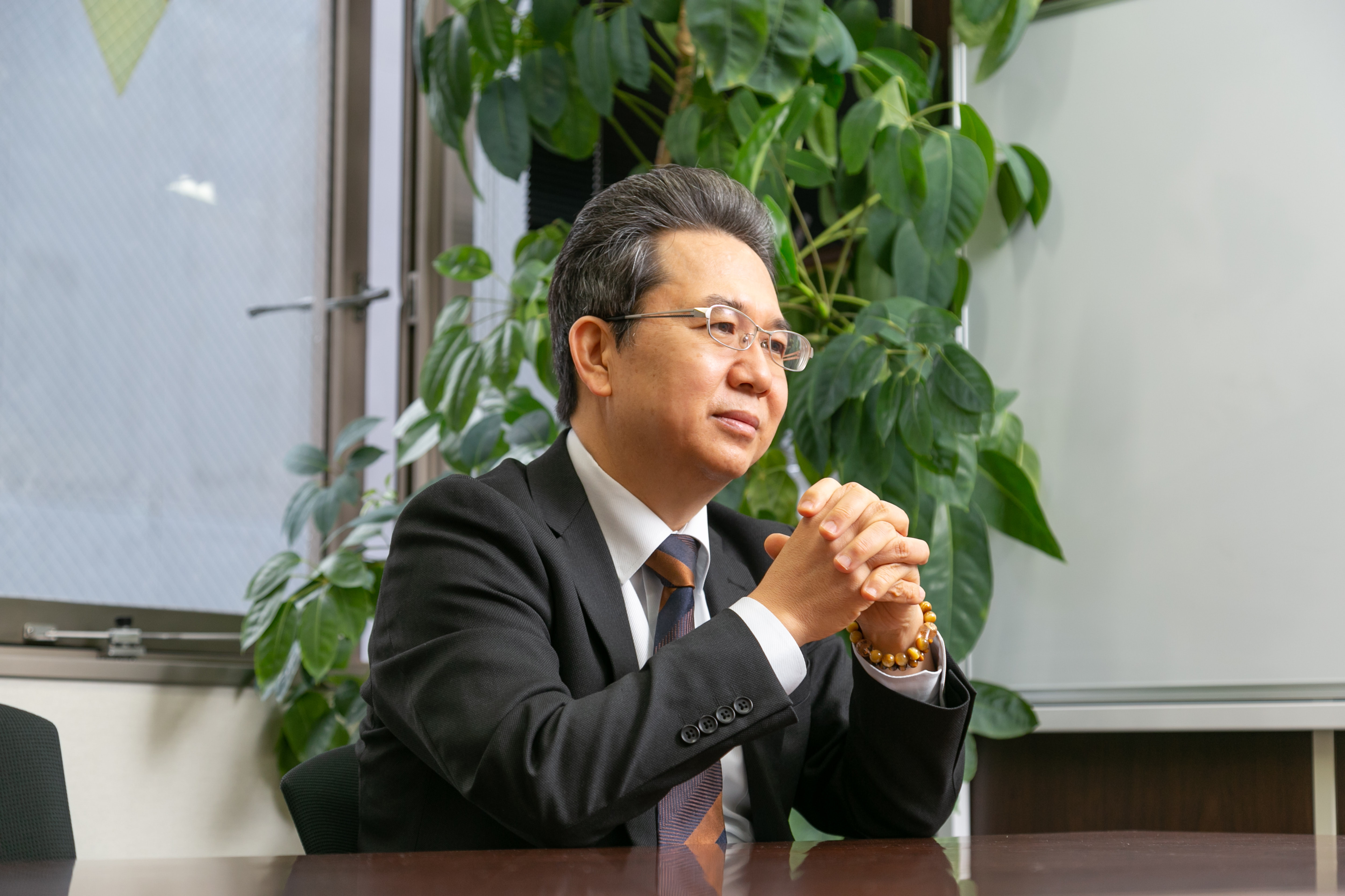 観葉植物を背に、両手を組んで穏やかな表情で語りかける、株式会社マネジメントオフィス・K代表取締役・川原一紀